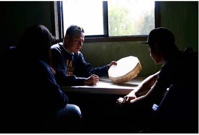 Photo: NAISC scholar mentoring youth with small drum at table near window.
