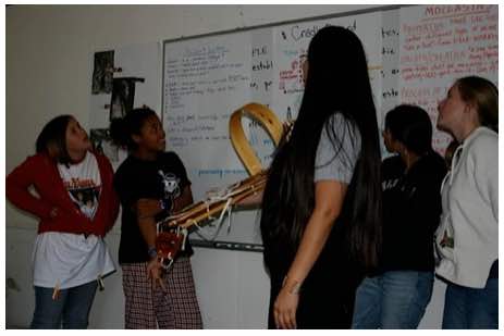 Photo: Elvina Charley holding cradle board standing with girls in classroom in front of white board with writing.