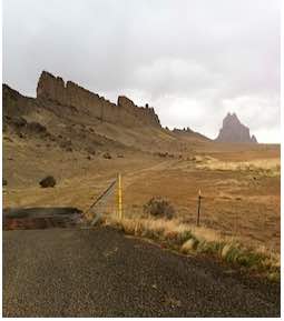 Photo: Shiprock scene with fence and road in foreground, mesa in background