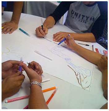 Photo: Students' hands and arms drawing on white paper on a table